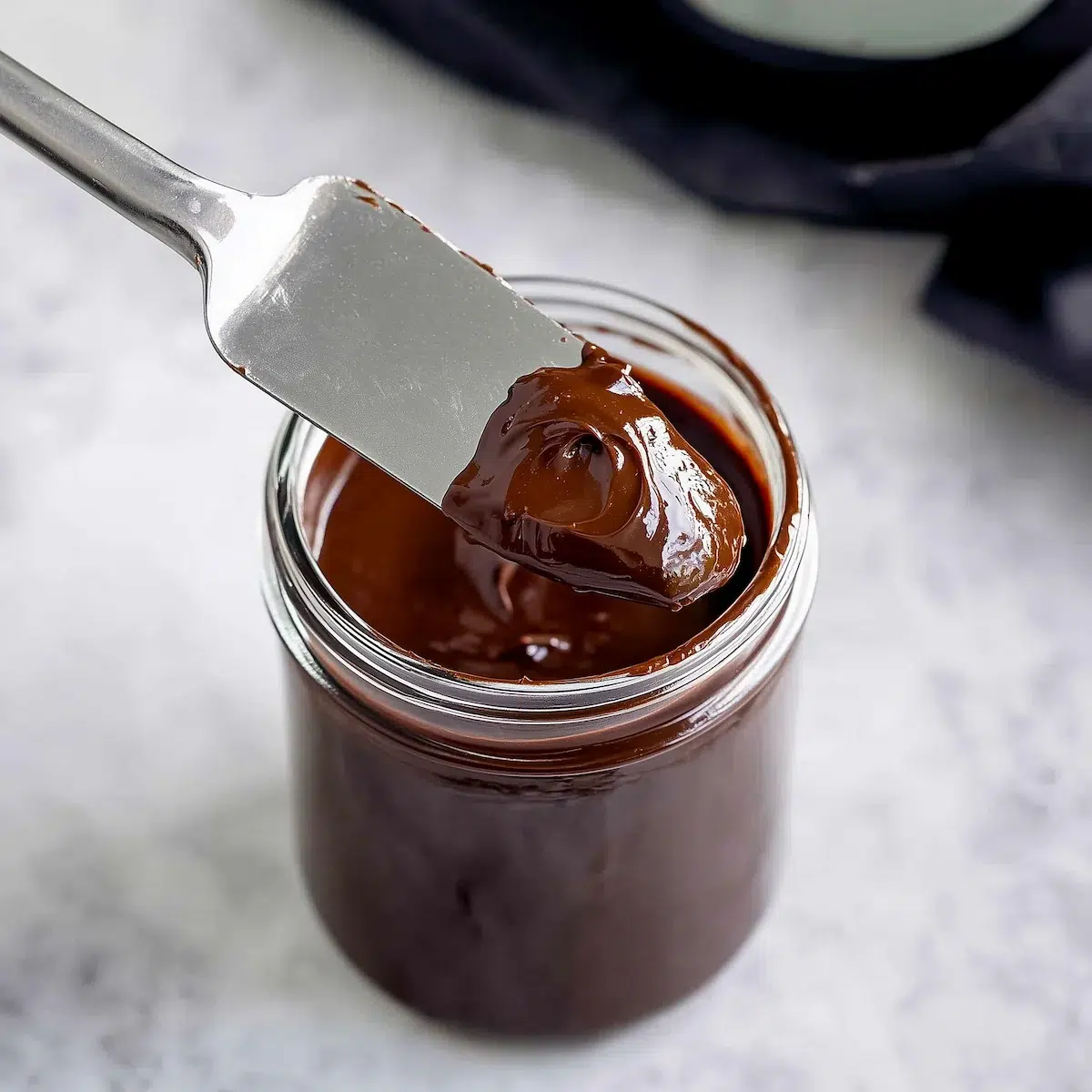 fudge-frosting-for-gluten-free-chocolate-zucchini-cake Glossy chocolate fudge frosting for gluten free chocolate zucchini cake in a glass jar with a metal spatula lifting a swirl of frosting